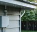 A weather-resistant junction box mounted on a shed wall under an overhang, surrounded by rain and snow, showcasing its durability and protective capabilities in a lush outdoor setting.