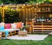 "Charming backyard entertainment setup featuring string lights, DIY pallet furniture with colorful cushions, and a rustic beverage station against a lush green backdrop."