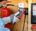 A person wearing OSHA-recommended protective gear, including insulated gloves and safety glasses, using a voltage tester on an electrical outlet inside a shed. Visible safety equipment includes a fire extinguisher and a clearly labeled circuit breaker panel.