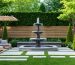 A beautifully landscaped backyard featuring a wooden acoustic fence, dense evergreen trees, and a tranquil tiered fountain, illustrating effective sound dampening solutions to create a peaceful outdoor space.
