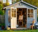 Small wooden 8x10 studio shed in a residential backyard at golden hour, door partially open to reveal an easel and organized art supplies, with house and trees softly blurred in the background.