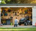Homeowner kneels to measure a riding mower while holding a tablet inside an open wooden storage shed with organized shelving, workbench, and garden tools; warm natural light and a softly blurred suburban yard beyond.