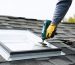 DIY homeowner installing a curb-mounted skylight on an asphalt-shingle shed roof with metal flashing and sealant arranged, photographed from slightly above under bright overcast light, with a garden and toolshed softly visible in the background.
