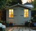 "A garden shed warmly lit from within during dusk, with a clear view of plants and trees in the background."