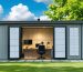 A serene backyard scene with a soundproofed shed converted into a peaceful workspace. Visible soundproofing features and a person working inside, surrounded by lush greenery and a clear sky.