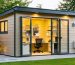 Modern modular backyard office shed with wood siding, open glass door revealing a desk and chair, and an exterior mini-split unit, photographed at golden hour with lawn, garden path, and house softly blurred in the background.