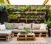 DIY pallet sofa with cushions and a pallet coffee table next to a vertical herb garden on a small backyard patio at golden hour, with fence and spare pallets softly blurred in the background.
