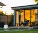 Modern backyard shed office with a person on a laptop, receiving signal from a white outdoor WiFi access point mounted under the house eave across a green lawn at golden hour.