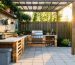 DIY outdoor kitchen on a small patio with a pallet countertop topped by concrete pavers, a charcoal kettle grill, a two-burner camp stove, storage bins underneath, and a simple pergola with bamboo shade at golden hour.