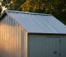 Close-up of a shed corner with standing seam metal roofing and metal flashing as glowing embers drift by, lit by warm golden-hour light, with a blurred gravel defensible space, distant trees, and a firewood stack stored far from the structure.