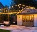 Backyard at dusk with a modern wooden shed illuminated by overhead string lights, mid-level wall sconces, and low path lights leading to the door, with patio seating and trees softly visible behind.