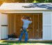 Person stapling reflective foil insulation to the interior roof of a wooden backyard shed on a sunny day, with green lawn and trees softly blurred in the background.