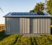 Eye-level view of a gray corrugated steel backyard shed on a gravel pad, framed by native plants, with solar panels on a nearby roof and a distant wind turbine in warm evening light.