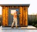 Homeowner using a drill to attach an interchangeable wall panel on a modular shed made of reclaimed wood with a recycled metal roof, with a garden, reclaimed boards, and removable shelving softly blurred behind.