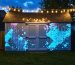 Twilight backyard scene with a weathered garden shed illuminated by projection-mapped geometric patterns and magenta/teal LED uplights, a warm string light sculpture spanning a pergola above, and softly blurred fence, plants, and tree silhouettes in the background