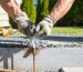 Gloved hands sealing a small gap around an exterior utility pipe with steel wool while holding a caulk gun at a house foundation; blurred background shows gravel border, trimmed shrubs, a lidded metal trash bin, and snap traps along a fence.