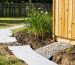 Eye-level wide shot of a wooden backyard shed beside a gravel-filled trench with a visible perforated pipe, soil graded away from the foundation, and a small rain garden of native plants in the background on a damp lawn.