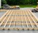 Pressure-treated shed floor frame with evenly spaced joists on concrete block piers in a backyard, photographed from a 45-degree angle under soft overcast light, with a blurred house, tools, and stacked boards in the background.
