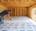 DIY homeowner kneeling in a wooden shed, fitting rigid foam insulation between exposed floor joists over a clear vapor barrier, with soft daylight from an open door and a blurred tool wall and garden in the background.