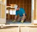 Homeowner kneeling in a wooden shed, placing rigid foam boards between exposed floor joists under an open floor section, with natural daylight from an open door and a blurred workbench and garden in the background