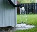 Corner of a wooden shed during rainfall with water pooling at the roof edge and draining into an aluminum gutter and downspout, flowing through a black extension to a gravel bed several feet from the foundation; wet siding, lawn, and a simple fence in the background.