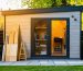 Modern backyard shed with wood-look composite cladding and subtle metal trim at golden hour, with a sawhorse holding various unlabeled siding samples in the foreground and a softly blurred garden and fence behind.