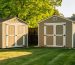 Oblique aerial view of a suburban backyard with two wooden storage sheds spaced several feet apart on a lawn, with house, driveway, garden beds, and trees in soft golden hour light.
