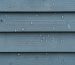 Close-up of a weatherproof 4x8 panel being screwed onto a timber-framed backyard shed, with water droplets on the surface and a blurred yard, tools, and stormy sky in the background