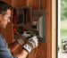DIY homeowner wearing safety glasses and insulated gloves wiring a transfer switch next to a main electrical panel on a shed wall, soft daylight with blurred tools and a portable generator in the background.