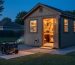 Workshop shed at dusk glowing with interior lights, portable generator placed safely on a pad about 20 feet away with exhaust directed away, heavy-duty cable connected to an exterior power inlet; trees and fence in the background.