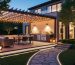 Modern backyard patio at dusk with overhead string lights, lanterns on tables, task lighting over a dining area and grill, and subtle accent lights along steps and a stone pathway with uplighting on a tree and pergola.
