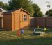 Wooden garden shed near a backyard fence with red, blue, and yellow utility flags and a green utility box marking an easement; a string line outlines the proposed footprint; neighboring house and power pole softly blurred in the background.