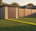 Backyard scene with a modern shed positioned several feet from a wooden fence marking the property line, a yellow measuring tape stretched on the grass between them, warm late-afternoon light, and a softly blurred house and trees in the background.