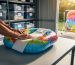 Hands rolling a colorful inflatable pool float with white tissue paper on a workbench in a clean storage shed, with clear sealed plastic bins on shelves and another inflatable drying in sunlight outside.