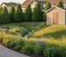 Slightly elevated view of a backyard with a curving rain garden and swale filled with native plants, a permeable paver path, and a small shed on raised ground, with a row of windbreak trees behind under warm evening light.