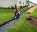 Homeowner kneels beside a gravel-filled french drain and grass swale carrying water away from a suburban house under overcast light, with a graded lawn, downspout extension, and straw erosion control blanket on a small slope in the background.