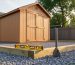 Low-angle photo of a 6x8 wooden shed on a compacted gravel pad with timber edging, a bubble level on the base, and a hand tamper nearby, with a blurred backyard fence and trees in the background.