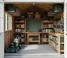 Eye-level view from the doorway into a compact 8x8 backyard shed with floor-to-ceiling shelves, pegboard tools, overhead racks, a small workbench, and a lawnmower, illuminated by soft natural light with a blurred garden visible outside.