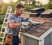 Person installing asphalt shingles on a small backyard shed roof from a slight low angle, wearing safety gloves and eye protection, with garden, ladder, and materials softly blurred in the background under diffused afternoon light.