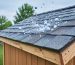 Close-up of impact-resistant asphalt shingles on a shed roof as golf-ball-sized hailstones hit, with a softly blurred backyard of trees and fencing under overcast light