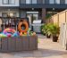 Two large collapsible outdoor storage containers on a small patio, one expanded with garden tools and pool toys, the other folded flat against a wooden fence, in soft late-afternoon light with a blurred townhouse and potted plants behind.