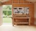 Organized shed interior with plywood and tongue-and-groove pine walls, pegboard tools above a wooden workbench, shelves, and natural side-window light, with a garden visible beyond the doorway.