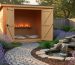 Small wooden garden shed converted into a meditation space, doors open to show floor cushions and a simple candle shelf, surrounded by lavender plantings, a river-rock path, and a bubbling water feature in warm early-morning light, with trees and a fence softly blurred behind.