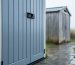 Close-up of a resin outdoor storage shed with water droplets beading on textured panels during light rain, with a rusty metal shed and a weathered wooden shed blurred in the background of a wet backyard.
