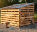 Backyard scene showing a small wooden pallet storage shed with a corrugated metal roof on a gravel foundation, with a drill, screws, and a lawn rake beside it in warm late-day light.