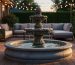 Tiered stone fountain with water flowing on a residential patio at dusk, surrounded by cushioned outdoor seating, potted plants, and warm string lights.