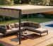 Modern outdoor sectional with built-in retractable canopy extended over seating on a sunlit deck, with landscaped garden, pool, and grill softly blurred in the background.
