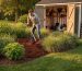 Homeowner spreading bulk mulch around a native plant bed beside a partially replaced lawn with ornamental grasses, with a tidy tool shed and stored mulch visible in the background at golden hour.