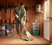 Homeowner in gloves sweeping spilled birdseed and cobwebs from a wooden shed floor, with sealed plastic storage bins neatly stacked, rakes hanging on the wall, clay pots raised off the floor, and daylight streaming through an open door.