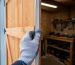 Gloved hand pressing adhesive weatherstripping onto a wooden shed door jamb, with a snowy yard blurred outside and a tidy tool bench visible inside.